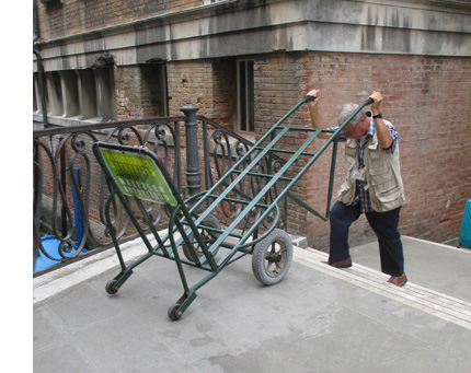 Photo shows a man with gray hair near the top of a bridge stairway, raising the handles of his cart up over his head.  The handles are at the ends of what looks like a ladder, and the other ends (at the front of the handtruck) each have a small wheel. The 'ladder' is resting on two large wheels near the front, and has a perpendicular support near the front to keep the luggage from sliding forward.